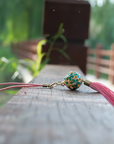 Zen Aroma Flora Bloom Sachet Pendant | Azure Bloom, featuring a golden floral filigree cage with green enamel blossoms and a pink silky tassel, displayed on a wooden surface with a blurred natural background.