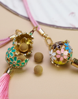Zen Aroma Flora Bloom Sachet Pendants with golden floral filigree cages and silky tassels, shown on a beige surface. One pendant is open, displaying aroma pellets inside.