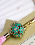 Zen Aroma Flora Bloom Sachet Pendant | Azure Bloom, featuring a golden filigree cage with green enamel floral blossoms and a pink silky tassel, displayed resting on a book.