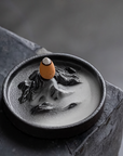 Ceramic incense burner with a small figurine on top, set against a dark background.