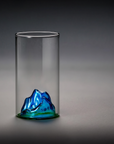 Cylindrical glass container with a blue and green mountain-like sculpture on a gray background