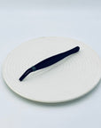 Zen Grip Tweezers resting on a ceramic white tray, showing the full matte black body and ergonomic ridged grip.