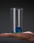 Hand holding a clear glass cylinder with a blue and green object inside against a dark background
