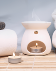 Zen Aroma unscented ghee candle burning inside a white ceramic Aroma Burner, with another tealight candle beside folded towels on a wooden surface — creating a clean, smokeless flame ideal for releasing aroma pellets.