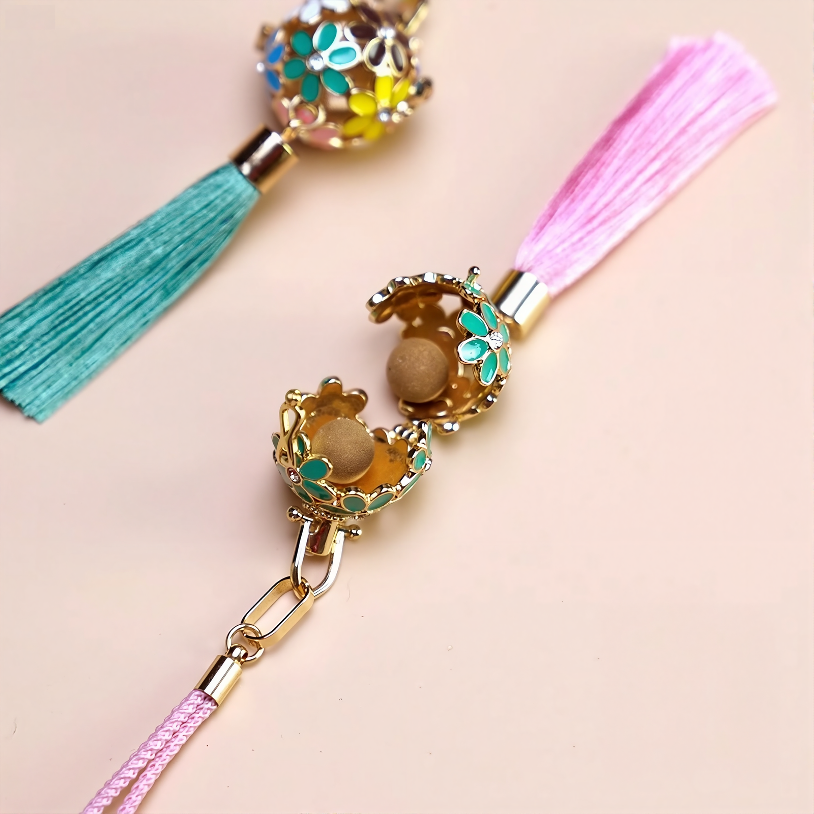 Zen Aroma Flora Bloom Sachet Pendant opened to reveal two aroma pellets inside its golden filigree floral cage, with green and pink silky tassels on a beige background.
