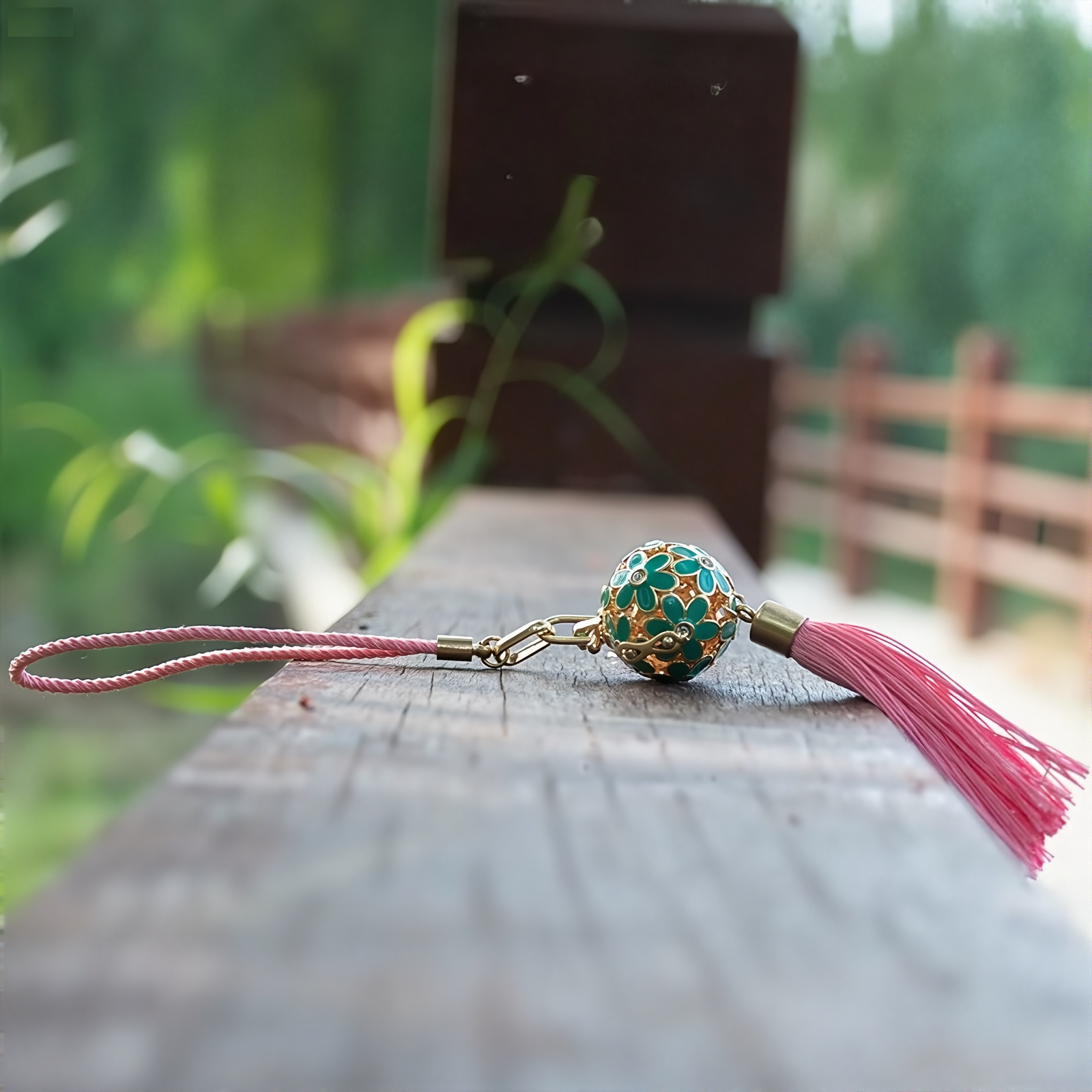 Zen Aroma Flora Bloom Sachet Pendant | Azure Bloom, featuring a golden floral filigree cage with green enamel blossoms and a pink silky tassel, displayed on a wooden surface with a blurred natural background.