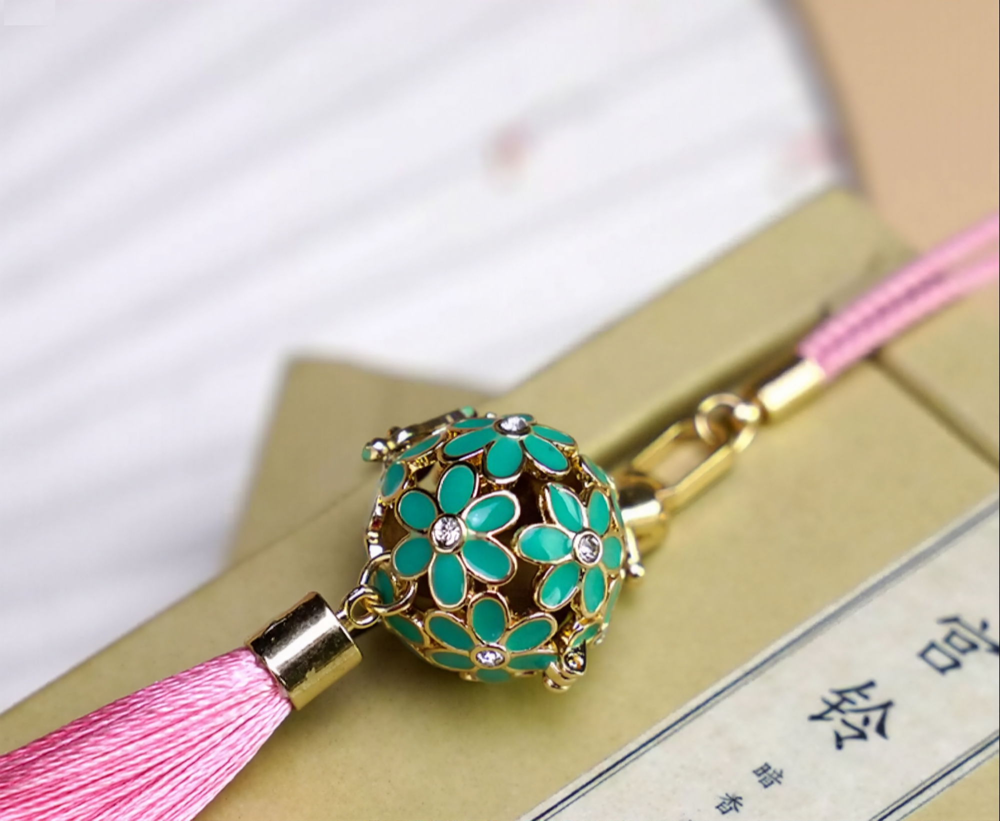Zen Aroma Flora Bloom Sachet Pendant | Azure Bloom, featuring a golden filigree cage with green enamel floral blossoms and a pink silky tassel, displayed resting on a book.