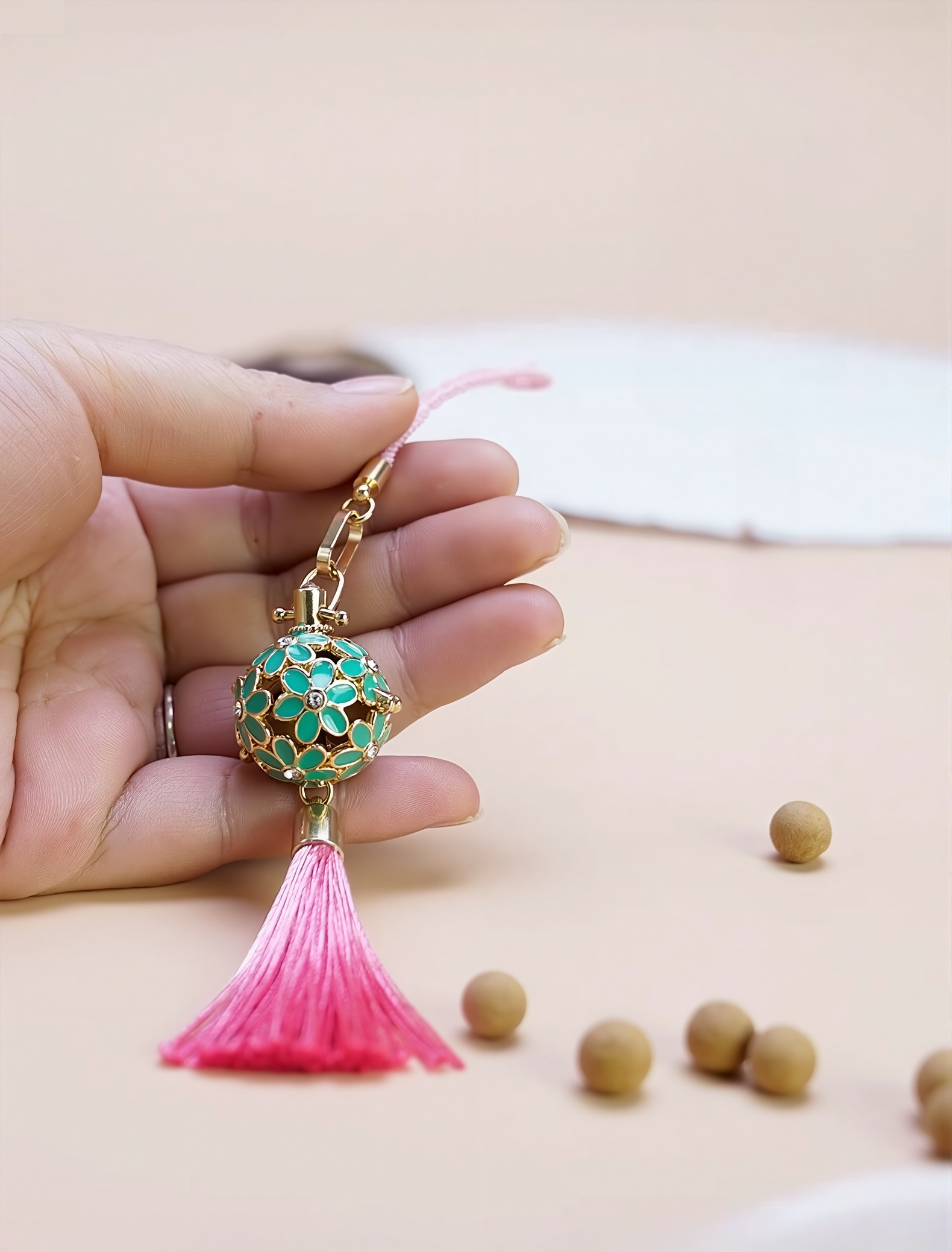 Hand holding the Zen Aroma Flora Bloom Sachet Pendant | Azure Bloom, showing its golden floral filigree cage with green enamel blossoms and a pink silky tassel, with aroma pellets scattered on a light background.