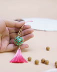 Hand holding the Zen Aroma Flora Bloom Sachet Pendant | Azure Bloom, showing its golden floral filigree cage with green enamel blossoms and a pink silky tassel, with aroma pellets scattered on a light background.