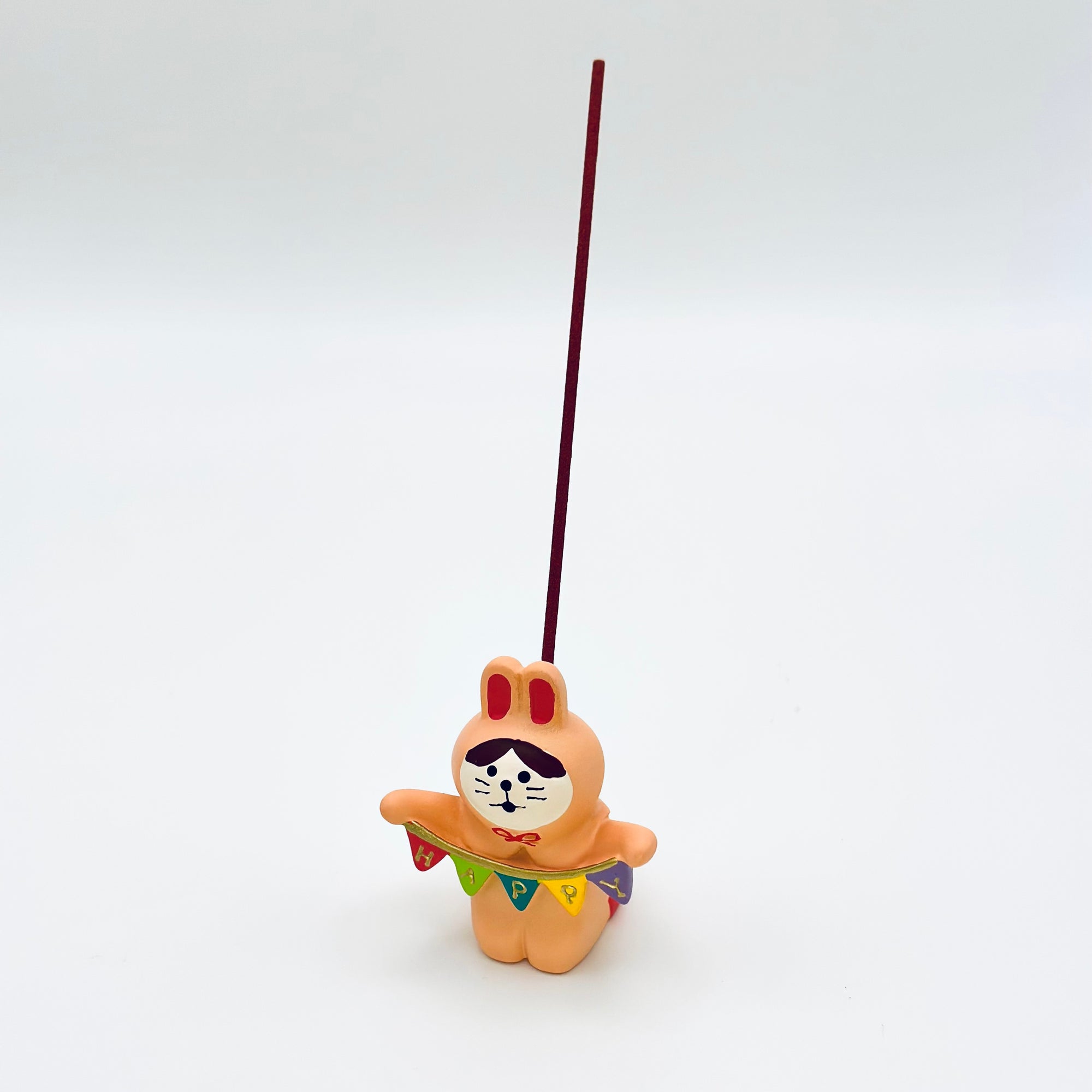Resin bunny incense holder in use, holding a lit incense stick upright behind a colorful “HAPPY” banner.