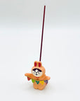 Resin bunny incense holder in use, holding a lit incense stick upright behind a colorful “HAPPY” banner.