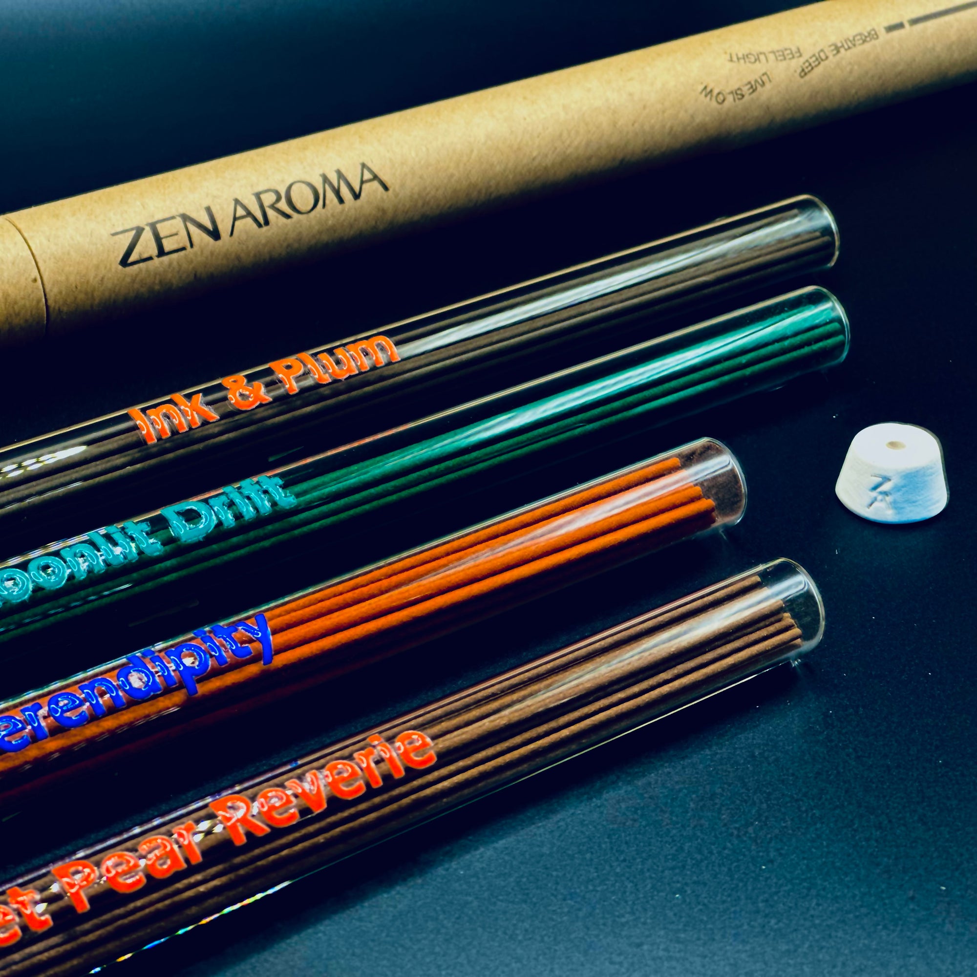 Zen Aroma incense collection close-up showing multiple glass tubes with colorful labels and natural incense sticks, displayed on a dark surface with brand packaging in the background.