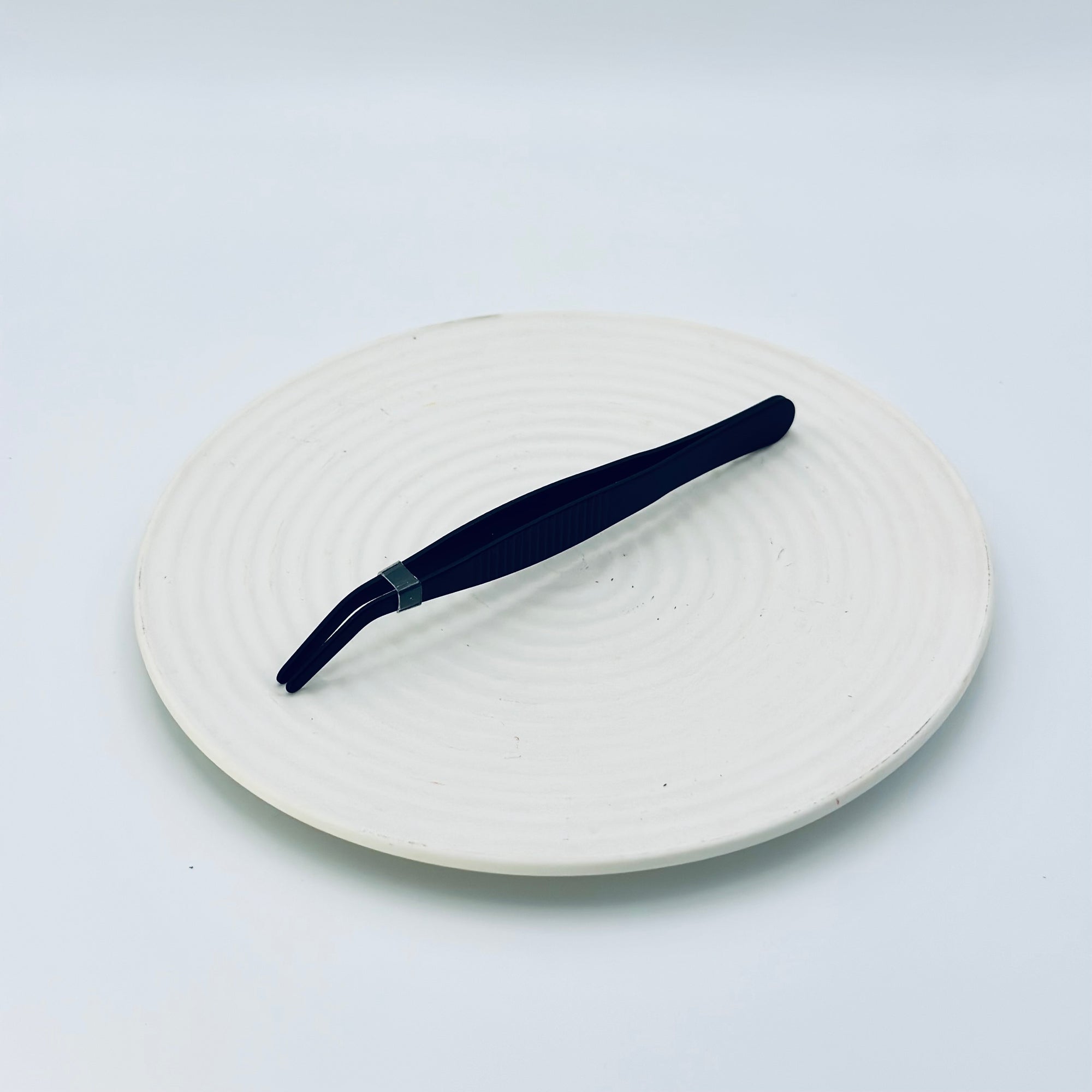 Zen Grip Tweezers resting on a ceramic white tray, showing the full matte black body and ergonomic ridged grip.