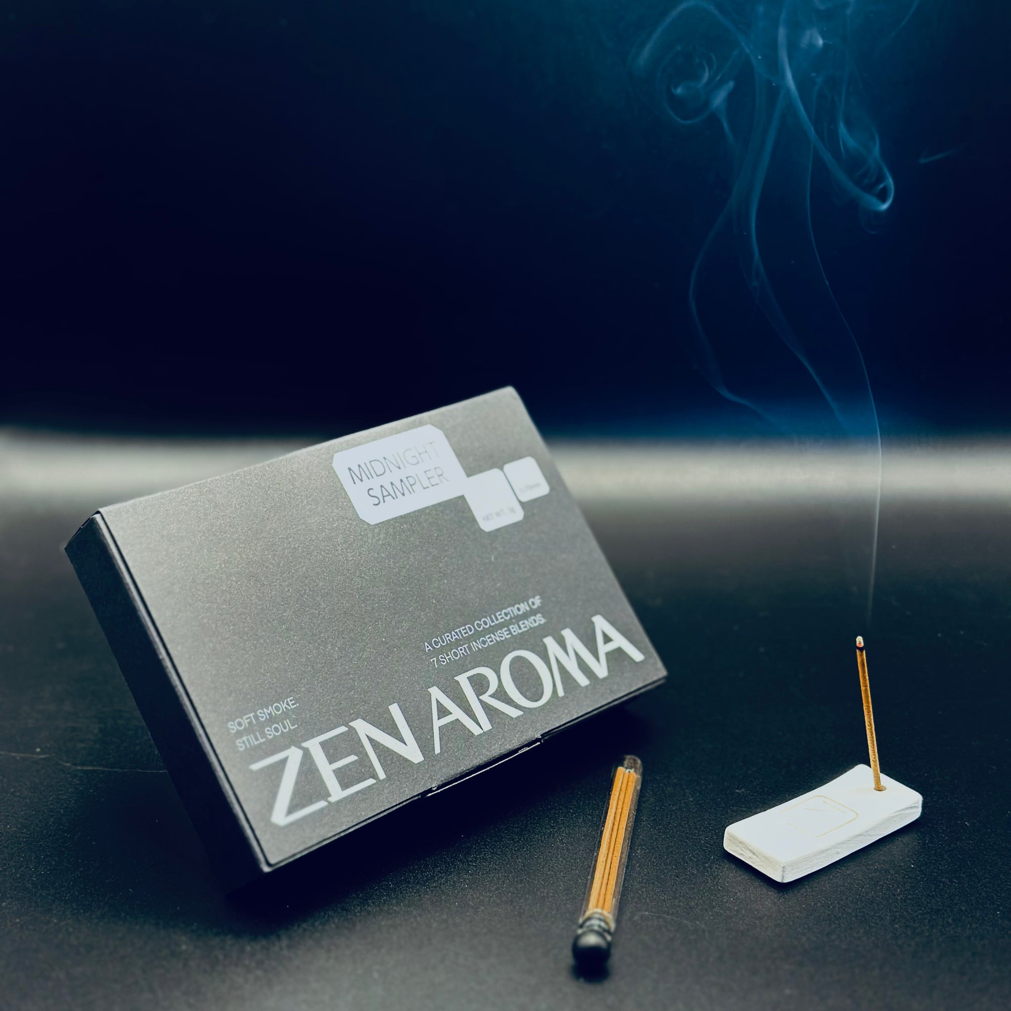 Zen Aroma “Midnight Sampler” box with a burning incense stick and gentle smoke rising in the background, styled on a dark surface to evoke a calm and elegant mood.