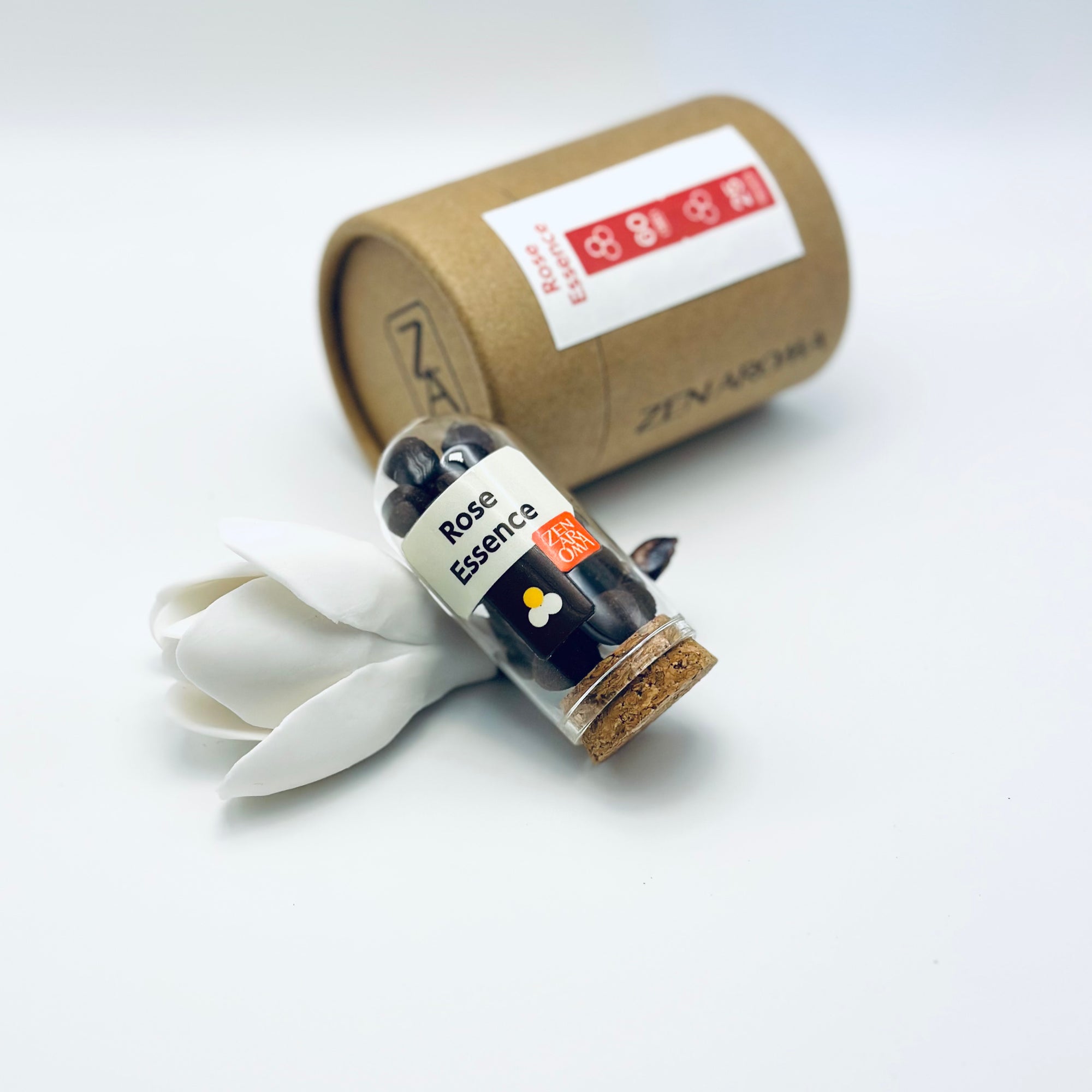 Zen Aroma Rose Essence aroma pellets in a glass jar with cork lid, displayed with a kraft paper tube and white flower petals on a light background.