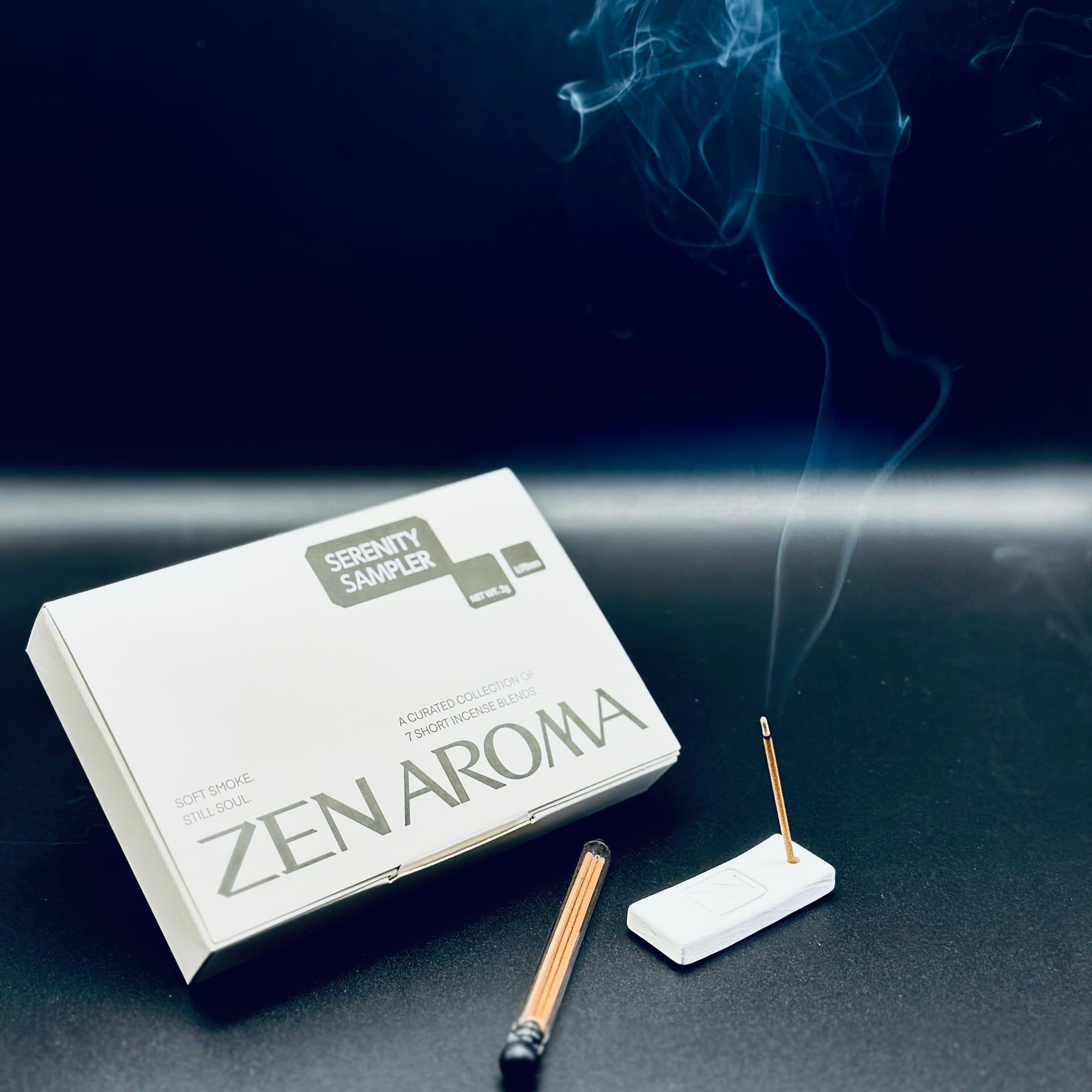 Zen Aroma “Serenity Sampler” incense set displayed with a burning incense stick releasing gentle smoke beside the box and holder, captured on a dark background to evoke calm and serenity.