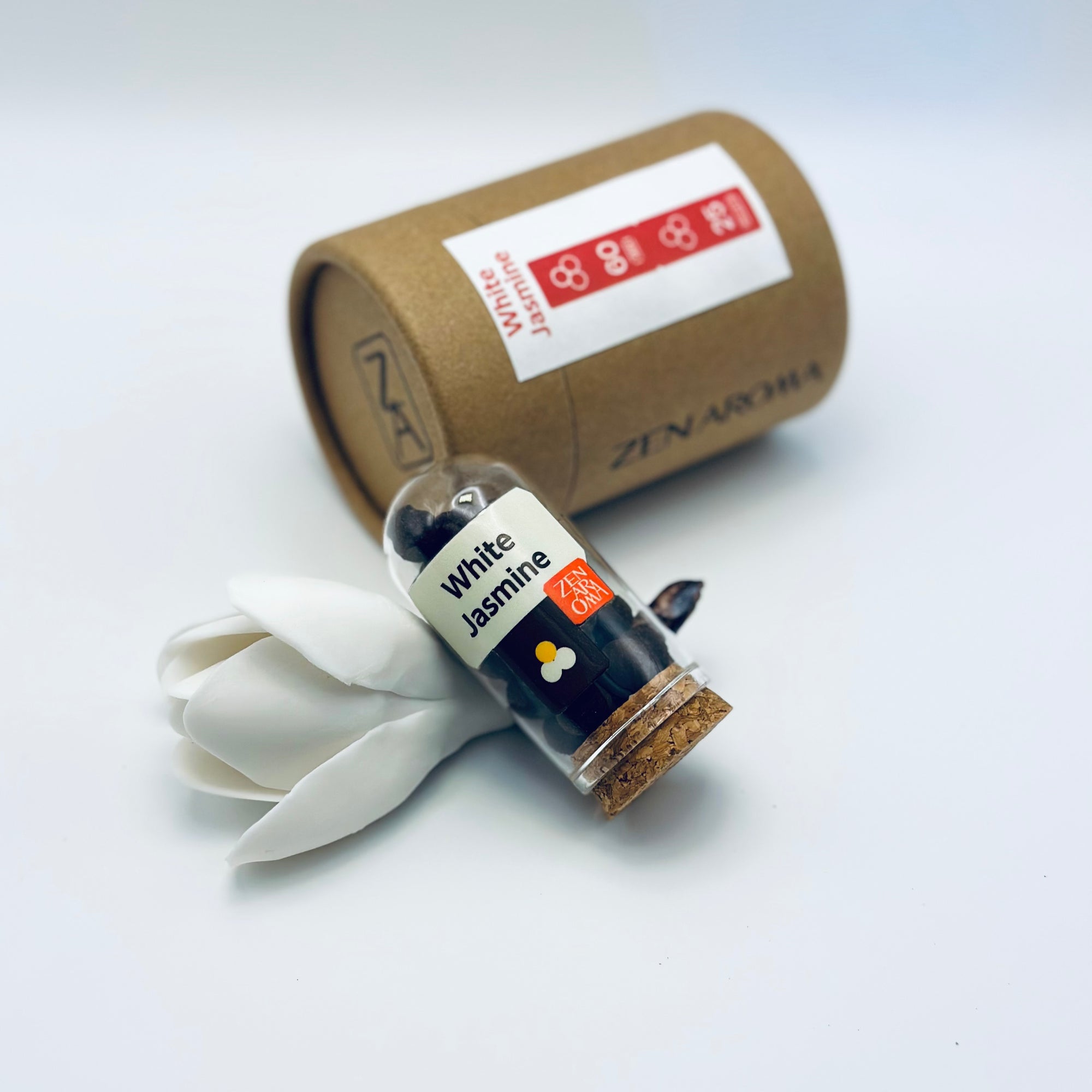 Zen Aroma White Jasmine aroma pellets in a glass jar with cork lid, displayed with a kraft paper tube and white flower petals on a light background.