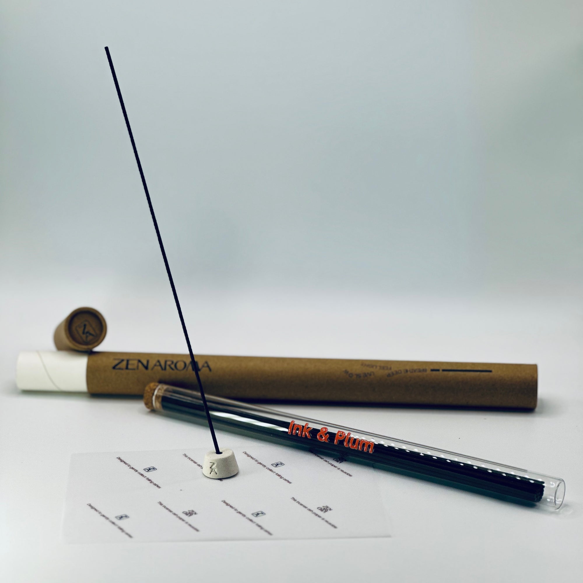 Zen Aroma Ink & Plum incense stick displayed with ceramic holder, glass tube, and brown cardboard tube packaging on a soft white background — minimalist and elegant home fragrance scene.